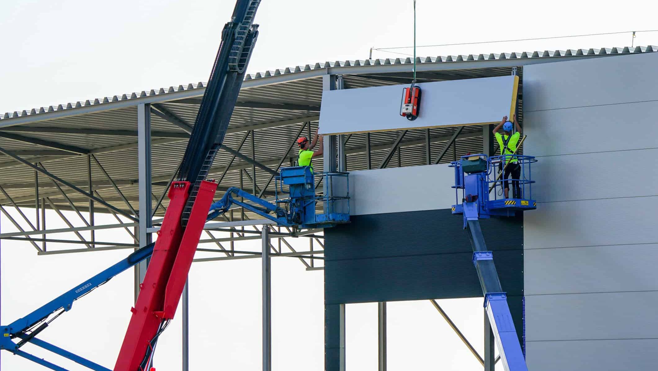 Arbeidere monterer fasadeelement på industribygg ved hjelp av kran og lifter under bygging.